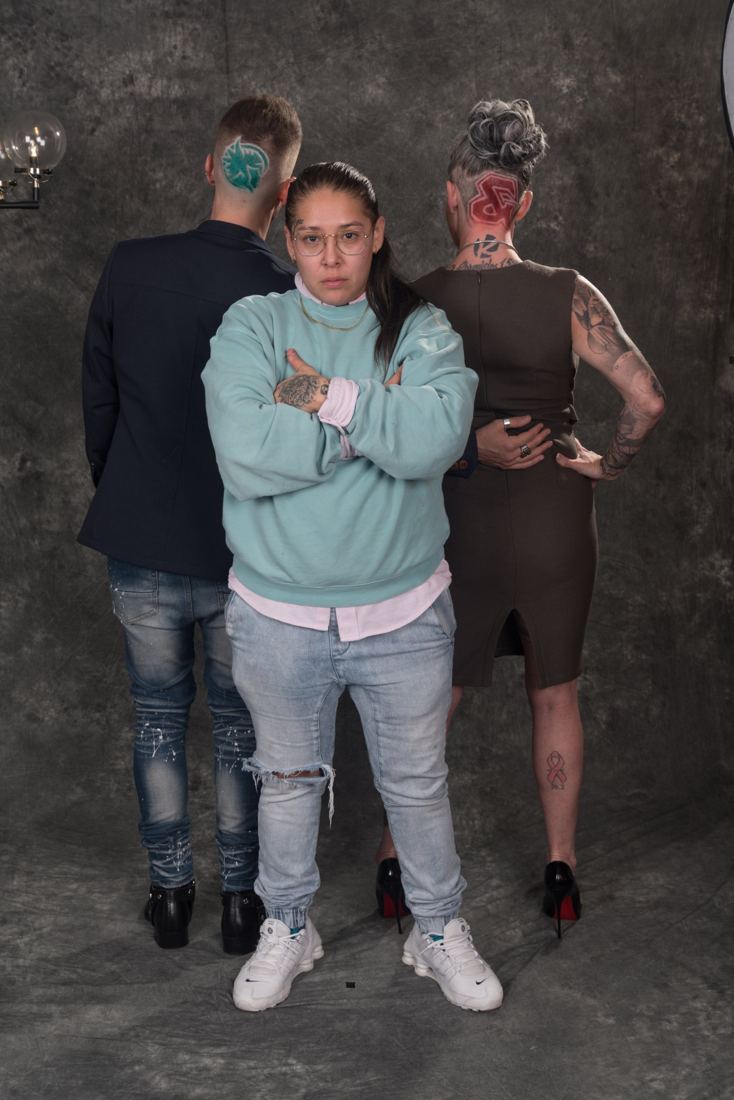 The barber standing front and center, arms crossed, with Carrie and Ian behind her showing their fresh cuts: a red ampersand and a teal logo