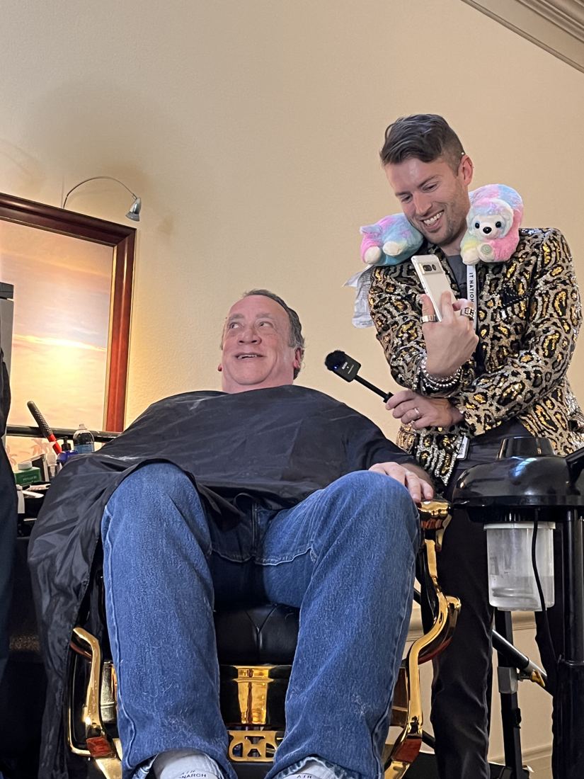 A friend in the barber chair on stage, caped and ready, with the host holding a mic